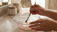 Woman mixing homemade face mask in glass bowl with wooden stick on marble bathroom counter surrounded by natural skincare products and cosmetic jars.