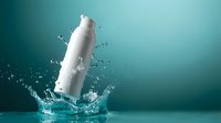 White cosmetic bottle falling into clear water creating dynamic splash against turquoise background for skincare and beauty product advertising.