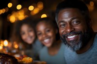Cheerful African American family enjoying dinner together with warm bokeh lights in background, father with grey beard smiling at camera during celebration meal.