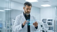 Middle-aged male scientist with salt-and-pepper beard wearing white lab coat examining holographic data display in modern laboratory setting. Research and innovation concept.