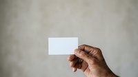 Person with medium-dark skin tone holding blank white business card against neutral gray textured background. Hand positioned for mockup or branding presentation use.