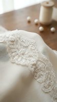 Cream linen fabric with delicate embroidered lace trim detail, styled on wooden table with pearl beads and ceramic vessel for elegant home decor and textile design projects.