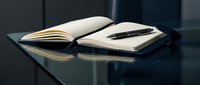 Open notebook with blank pages and black pen on dark reflective desk surface, suitable for business, education, writing, planning, and office concepts.