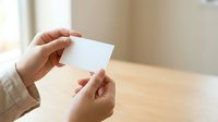 Woman holding blank white business card in both hands over wooden table with natural window light, suitable for corporate branding and identity mockups.