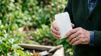 Senior man with weathered hands holding blank white card outdoors in garden setting with green foliage background, suitable for greeting cards and marketing materials.