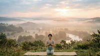 Woman in meditation pose sitting on wooden platform overlooking misty valley landscape at sunrise, wellness and mindfulness practice in nature.