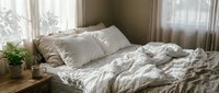 Cozy unmade bed with rumpled white linen bedding in sunlit bedroom corner with plant and coffee cup on wooden nightstand creating peaceful morning atmosphere.