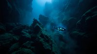 Deep underwater cave exploration with scuba diver navigating through rocky formations illuminated by bright blue sunlight filtering from above.