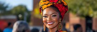 Young African woman wearing colorful traditional headwrap and gold earrings smiling warmly outdoors with natural lighting and blurred background.