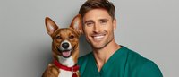 Young male veterinarian in green medical scrubs smiling with happy brown and white mixed breed dog wearing red collar against gray studio background.