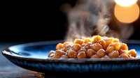 Steaming chickpeas served in blue ceramic bowl against dark background with golden bokeh light. Fresh cooked legumes with rising steam create appetizing mood.