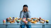 Young Asian man in blue shirt feeling overwhelmed surrounded by colorful donuts and sweets on table expressing stress from unhealthy eating habits and sugar addiction.
