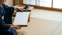 Adult male in traditional dark blue kimono holding blank clipboard with wooden frame in minimalist Japanese room with tatami mat flooring, suitable for business, education, and cultural content.