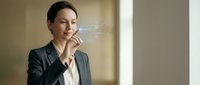Professional businesswoman in elegant suit enjoying morning coffee break in modern office with natural sunlight streaming through windows.