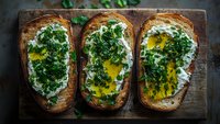 Three rustic sourdough toast slices topped with creamy ricotta cheese, runny golden egg yolks and fresh green herbs on wooden cutting board for breakfast brunch menu.