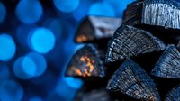 Stacked black charcoal firewood pieces with blue bokeh lights background creating moody atmosphere for winter and holiday season warmth concept.