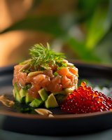 Fresh salmon tartare layered with diced avocado, garnished with dill and pine nuts, served alongside bright red caviar on black plate, elegant restaurant presentation.
