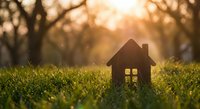 Small wooden house model on green grass with warm golden sunset light filtering through blurred trees in background creating dreamy bokeh effect for real estate concepts.