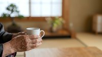 Senior man with wrinkled hands holding white ceramic mug with hot beverage in cozy home interior with blurred background and plants.