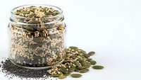 Mixed seeds and nuts in glass jar with scattered pumpkin seeds and chia seeds on white background for healthy snacking and cooking recipes.