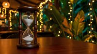 Vintage hourglass with flowing sand on wooden table surrounded by warm golden bokeh lights and tropical plants creating cozy atmospheric home decor ambiance.