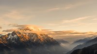 Golden sunrise illuminating snow-capped mountain peaks with misty valleys below, creating dramatic alpine landscape with warm light and atmospheric layers.
