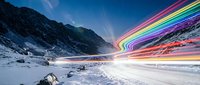 Snowy mountain landscape with vibrant rainbow light trails streaking across winter sky, creating dynamic motion effect over frozen terrain.