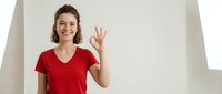 Happy young Caucasian woman in red v-neck t-shirt showing OK hand gesture with fingers, smiling at camera in bright modern interior space for positive communication.
