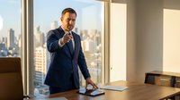 Confident businessman in navy suit pointing forward in modern office with city skyline view, leadership and success concept for corporate presentations.