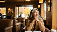 Thoughtful mature woman with gray hair sitting in cozy cafe restaurant, contemplating life decisions while enjoying warm atmosphere and natural lighting.