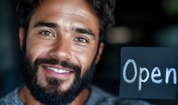 Smiling Hispanic male business owner holding open sign, showing genuine warmth and welcoming expression. Close up portrait with soft bokeh background.