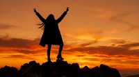 Woman silhouette with raised arms celebrating freedom on rocky cliff against dramatic orange sunset sky. Success achievement motivation concept.