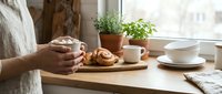 Woman enjoying morning coffee with fresh pastries on wooden table by sunny window creating cozy breakfast atmosphere at home.