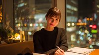 Young professional woman working late at office desk with city lights background. Business person in cozy workspace atmosphere for corporate lifestyle content.
