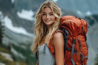 Young Caucasian woman with long blonde hair smiling while hiking in mountains, wearing gray tank top and carrying large orange backpack against blurred nature background.