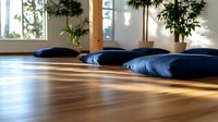 Meditation cushions arranged in row on wooden floor with natural sunlight streaming through windows, creating peaceful zen atmosphere in minimalist yoga studio.