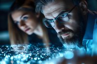 Focused bearded man with glasses analyzing glowing data visualization while female colleague works in background at modern tech office.