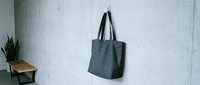 Gray canvas tote bag hanging on white wall in minimalist interior with wooden bench and plant for eco-friendly shopping and sustainable lifestyle.