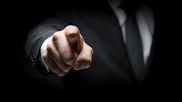 Businessman in dark suit pointing finger directly at viewer against black background, conveying authority and leadership in corporate setting.