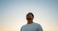 Man in light blue t-shirt looking upward against clear sky at sunset or sunrise, expressing hope, contemplation, or aspirational thinking for motivational and lifestyle content.