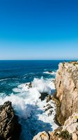 Dramatic coastal cliff with crashing waves and clear blue sky, featuring rocky shoreline and ocean spray for travel, nature, and landscape design projects.