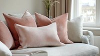Cozy sofa corner with soft pink velvet and cream textured pillows arranged on white upholstery, natural light from window creates serene living room atmosphere.
