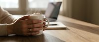 Professional woman holding white coffee mug at wooden desk with laptop in bright modern office workspace for remote work and business productivity.