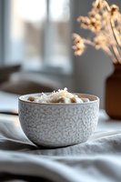 Creamy hot chocolate topped with coconut flakes served in textured white ceramic bowl on linen tablecloth, soft window light creates cozy atmosphere.