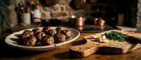 Homemade meatballs on white plate with fresh herbs on wooden cutting board in rustic kitchen setting with warm candlelight creating cozy dining atmosphere.
