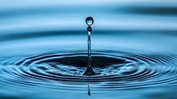 Water drop splash creating concentric ripples on blue surface with suspended droplet above, captured in high-speed macro photography showing liquid dynamics and surface tension.
