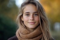 Young Caucasian woman with natural makeup and gentle smile wearing cozy brown knit scarf outdoors in autumn. Soft bokeh background creates warm atmosphere.