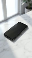 Modern smartphone with blank black screen lying on white marble surface near bright window with natural sunlight and green plant in background.