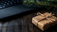 Small gift wrapped in rustic burlap with twine bow on dark wooden table with pine branch and laptop, creating cozy holiday workspace atmosphere.
