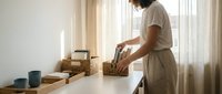 Young Asian woman organizing kitchen items in wicker baskets on white counter in bright modern home interior with natural lighting and minimalist decor.